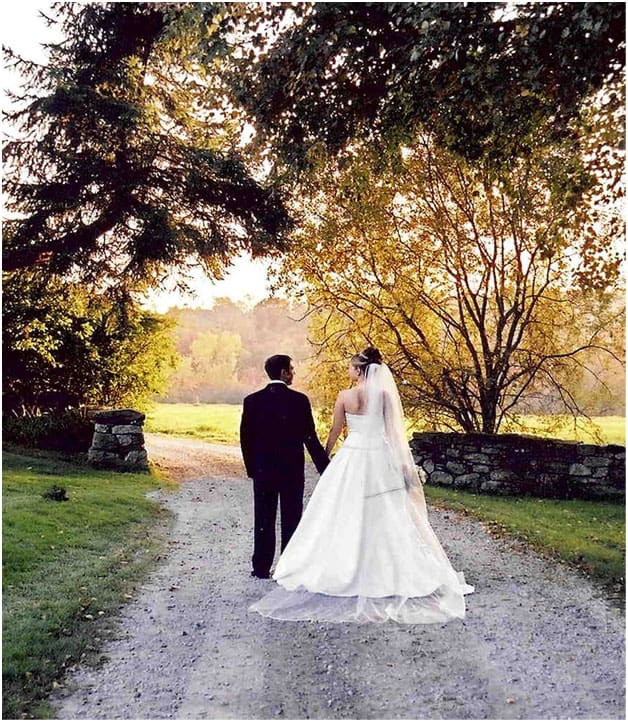 A bride wearing a white gown with her new husband in a black tux. Standing in the gravel driveway looking through a stone wall during early sunset