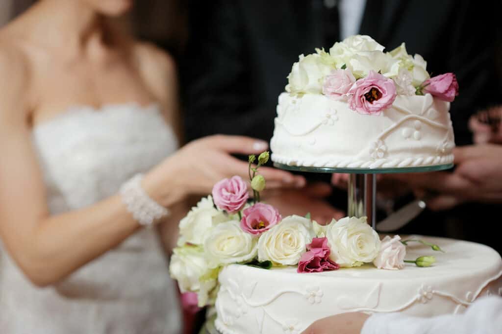 A white iced wedding cake with fresh white and pink roses as decoration. The bride in white appears in the background