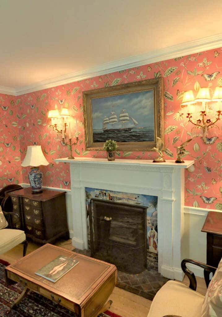 A white mantle around a fireplace with painted tiles. Pink wallpaper with various fauna and butterflies. A large ship painting above the mantle.