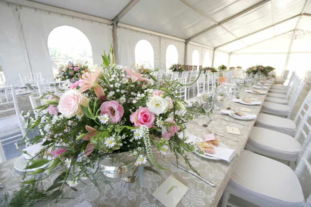 A long table decorated for a wedding in a tent. Lots of white chairs with beautiful bouquets of pink and white flowers.
