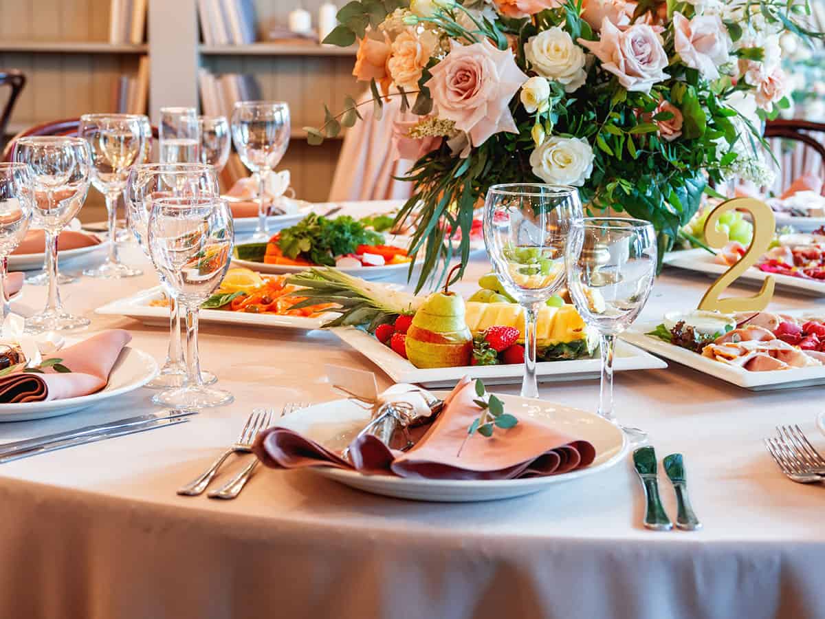 A closeup of a round table with each setting comprised of 2 stemmed glasses, white serving dishes and plates and a center piece of roses