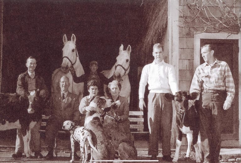 6 family members sitting in front of a barn. 2 white horses being held by one man, 2 calves, 2 Great Danes and 1 Dalmatian dog posed with the Richardson family.