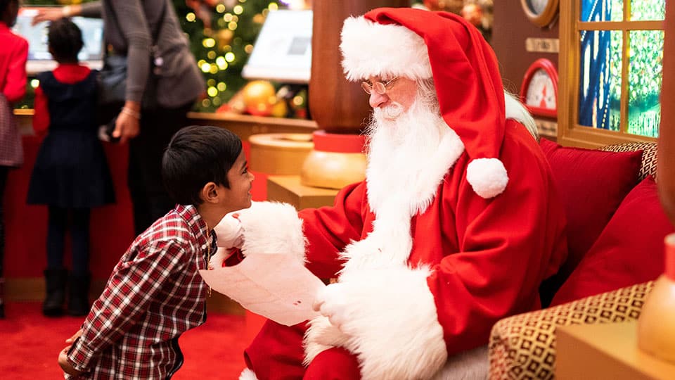 A white bearded Santa sitting down holding a Christmas wish list in his hands while a little boy looks on.